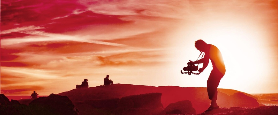 Ein Mann steht in einer Lavalandschaft und hält eine mittelgroße Filmkamera in Händen, die er auf zwei Personen am Horizont richtet. Das Bild ist insgesamt rötlich eingefärbt. 